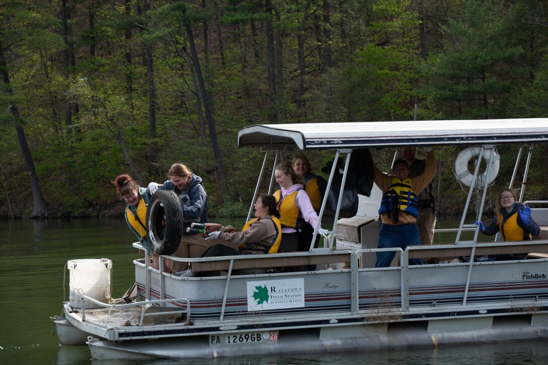 Individuals sitting on a pontoon boat on a lake lift a loose car tire from the water while collecting trash. Others sit under the boat's awning. The lake shoreline with trees are visible in the background.