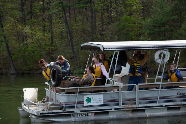Individuals sitting on a pontoon boat on a lake lift a loose car tire from the water while collecting trash. Others sit under the boat's awning. The lake shoreline with trees are visible in the background.