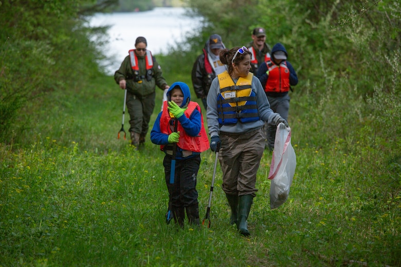 Individuals in life jackets carrying trash bags walk along a green grass trail collecting trash. The lake is visible on the horizon.