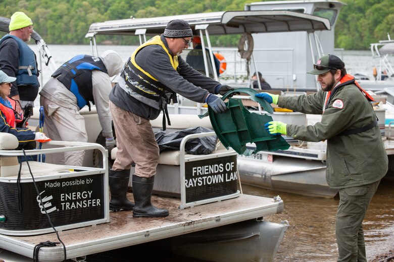 Two individuals, one standing on a boat and one standing on the shoreline, pass garbage for disposal.