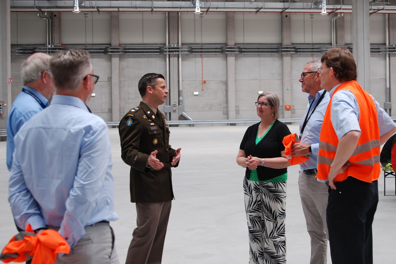 A group of people stand talking inside a large warehouse, one is wearing a dress military uniform.