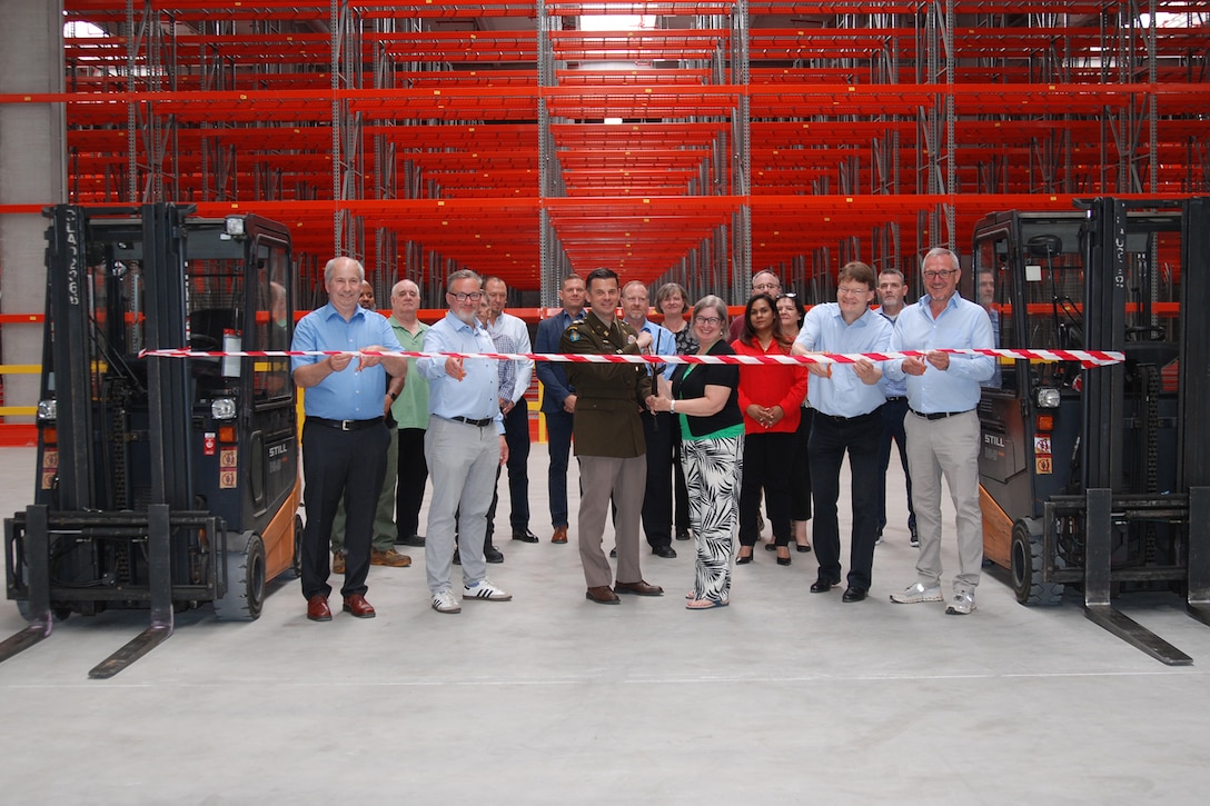A group of people stand in front of a ribbon suspended between two fork lifts inside a large warehouse, one is wearing a dress military uniform.