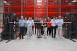 A group of people stand in front of a ribbon suspended between two fork lifts inside a large warehouse, one is wearing a dress military uniform.