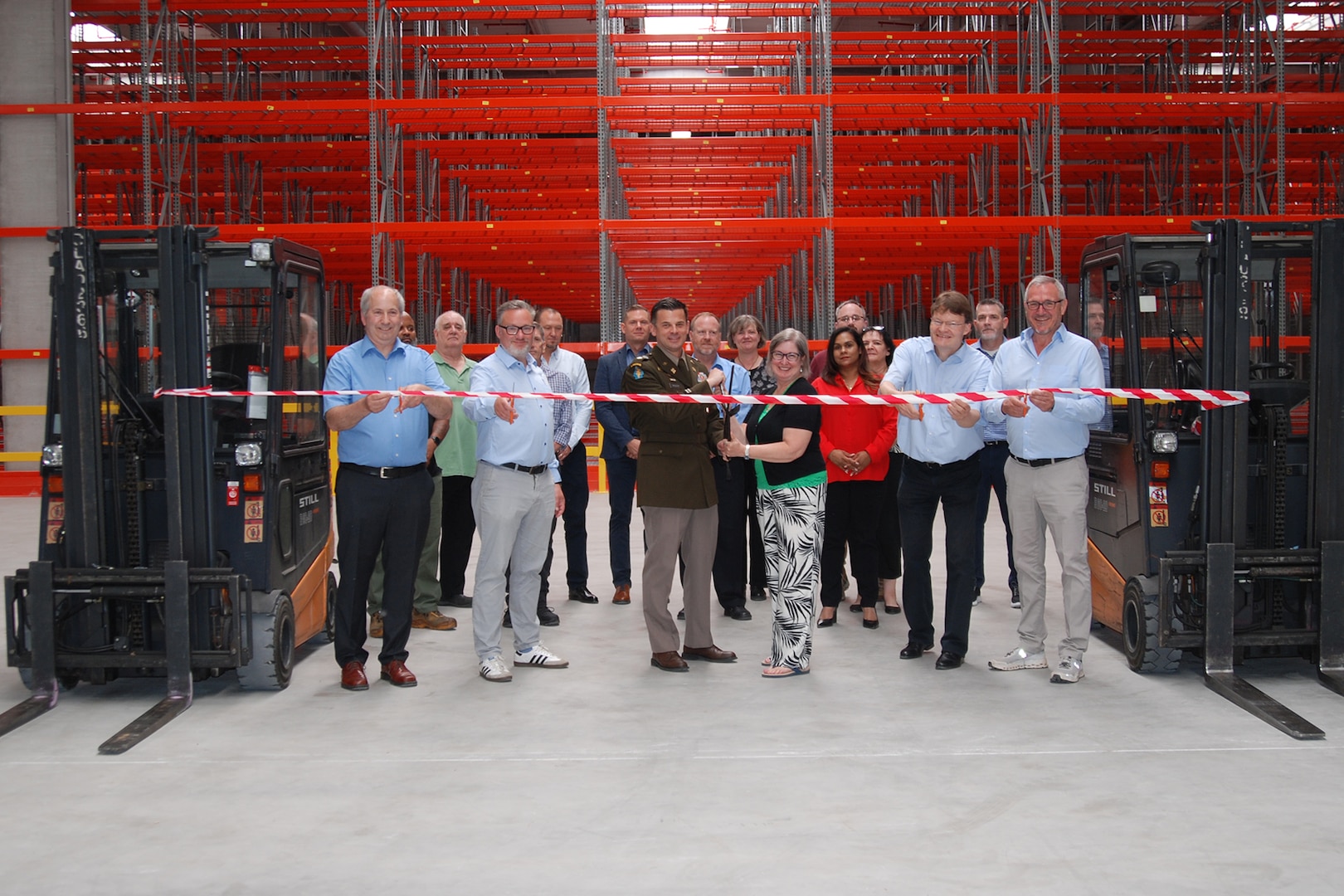 A group of people stand in front of a ribbon suspended between two fork lifts inside a large warehouse, one is wearing a dress military uniform.