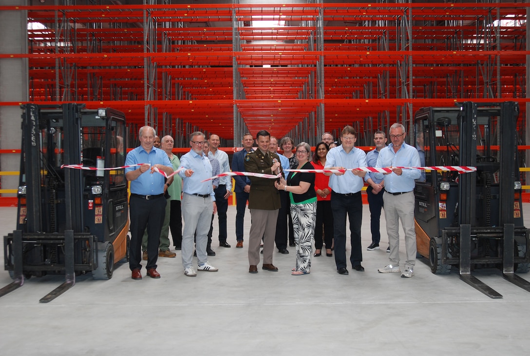 A group of people stand in front of a ribbon suspended between two fork lifts inside a large warehouse, one is wearing a dress military uniform.