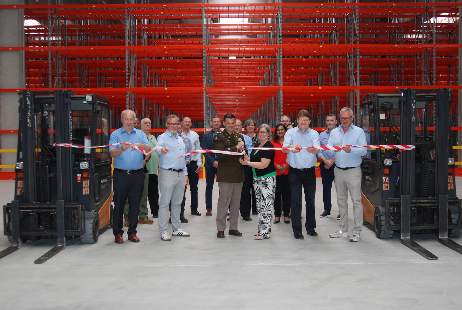 A group of people stand in front of a ribbon suspended between two fork lifts inside a large warehouse, one is wearing a dress military uniform.