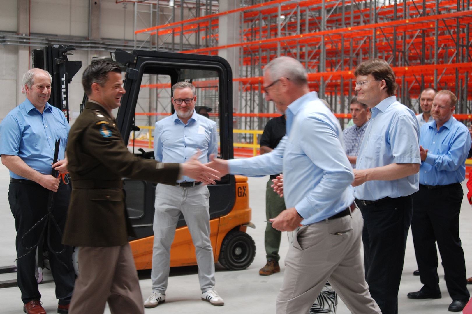 A group of people stand around talking inside a large warehouse, one is wearing a dress military uniform.