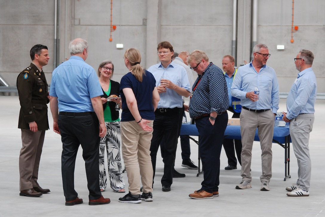 A group of people stand around talking inside a large warehouse, one is wearing a dress military uniform.
