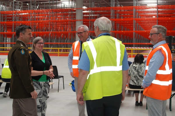 A group of people stand around talking inside a large warehouse, one is wearing a dress military uniform.