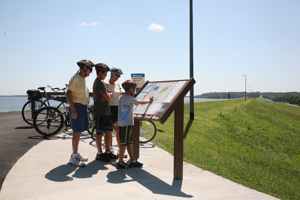 A family of four wearing bike helmets looks at posted signage between a lake and a grassy area, with bikes parked behind them.