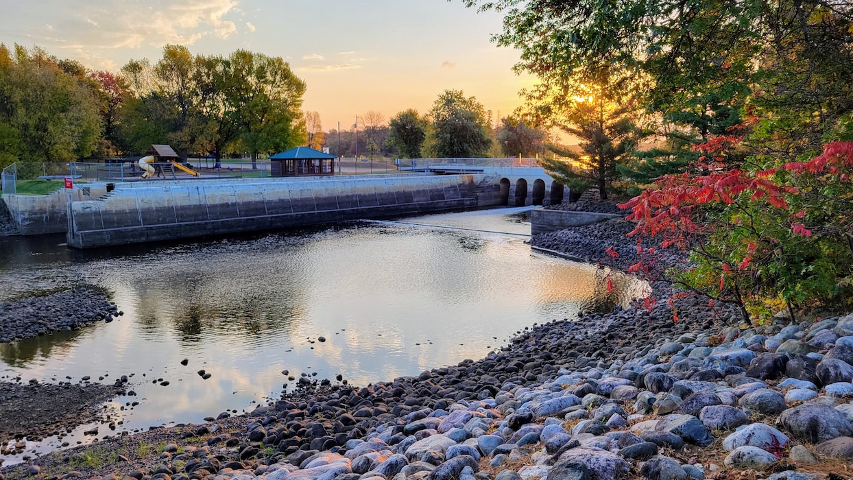 Photo of a river with a rocky forested shore leading to a dam at sunset.