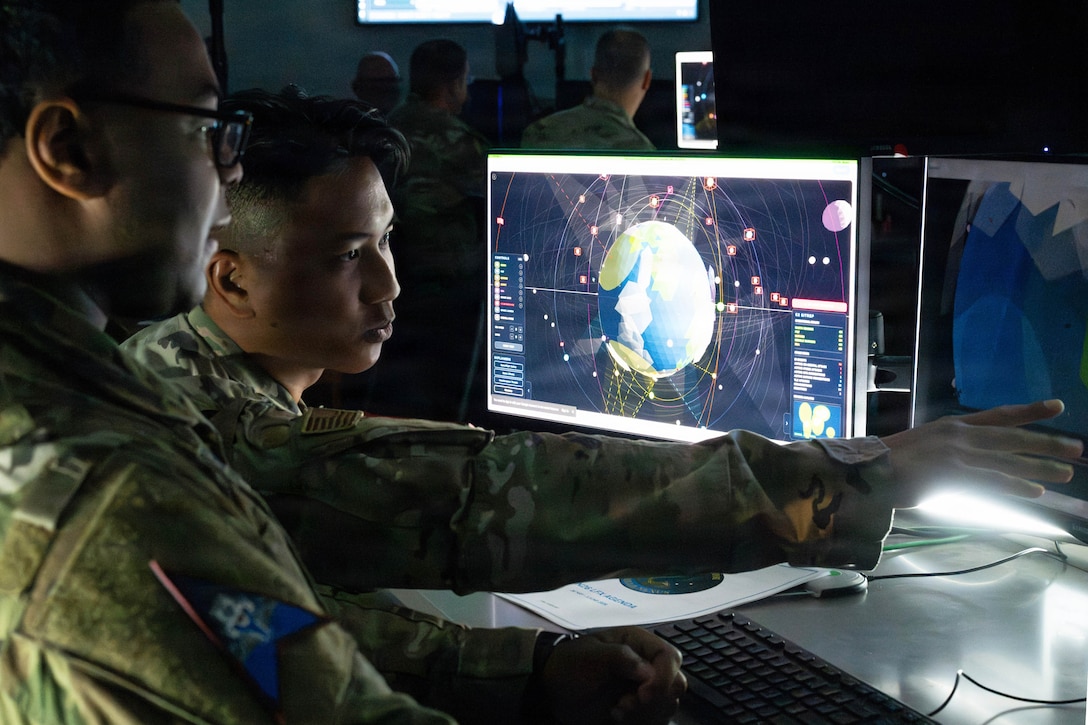Two guardians look at a monitor with Earth in space in a dimly lit operations center.