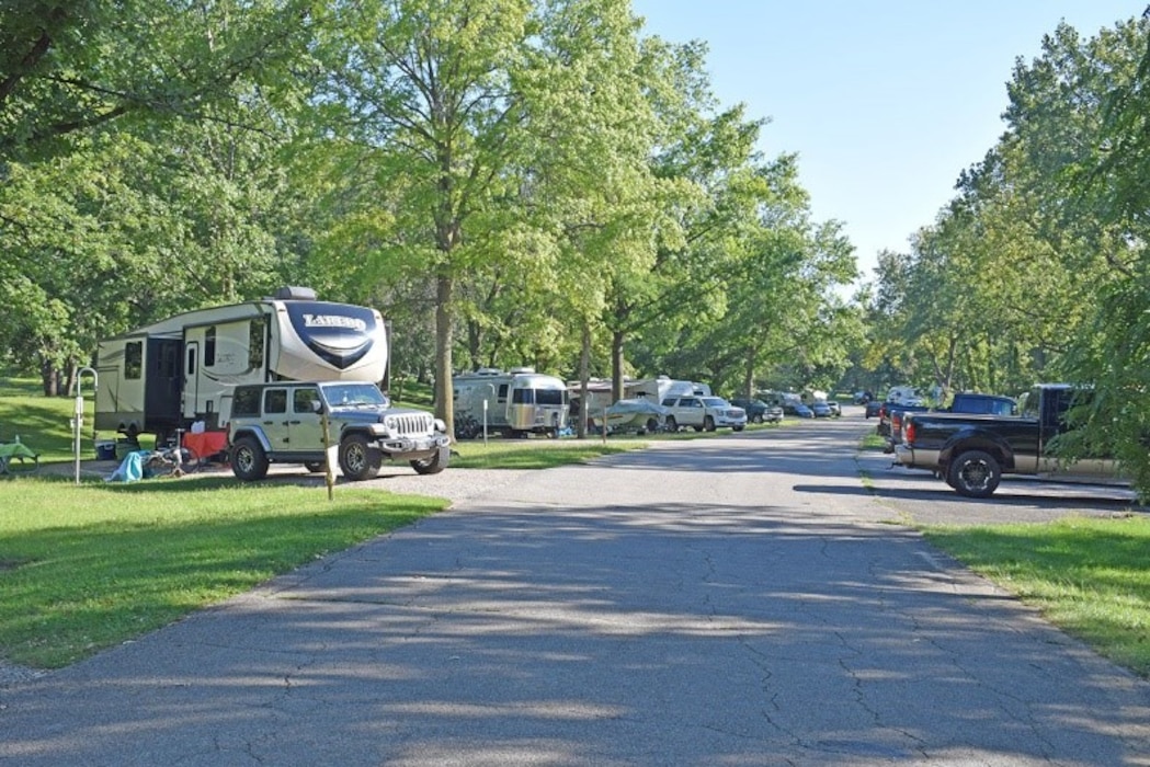 Multiple cars and RVs are parked along a wooded road.