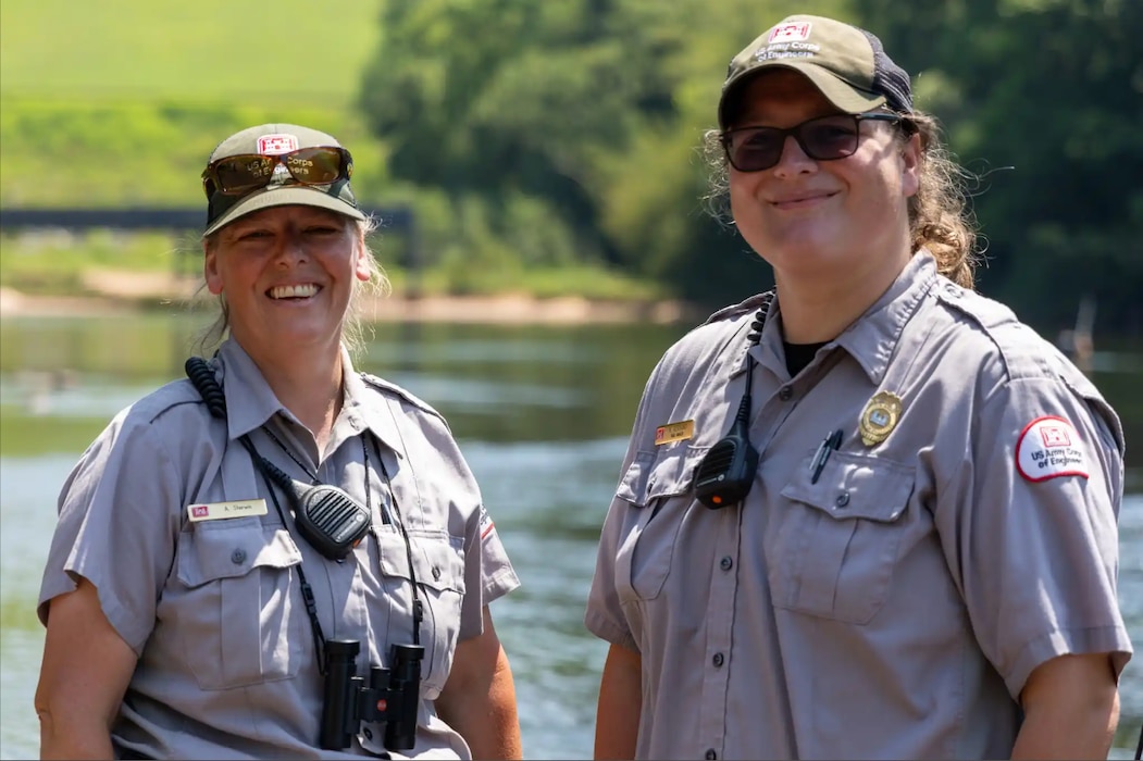 Two women wearing park ranger shirts, hats, and sunglasses smile at the camera.