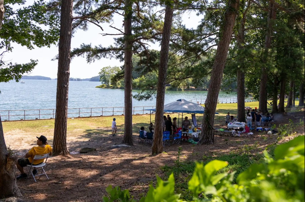 A large lake stands in the background of a wooded area under which people have set up a tent and chairs.