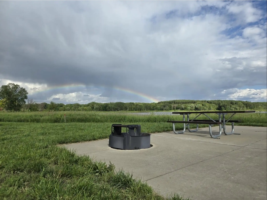 A concrete area with a grill and picnic table, next to a grassy area, with clouds and a rainbow in the background.