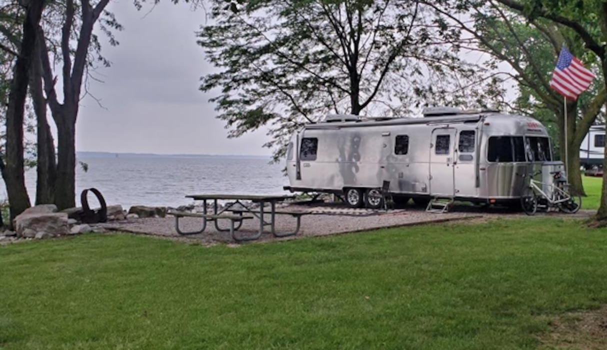 A large RV is parked next to a picnic table on a concrete pad between a grassy area and a lake behind it.