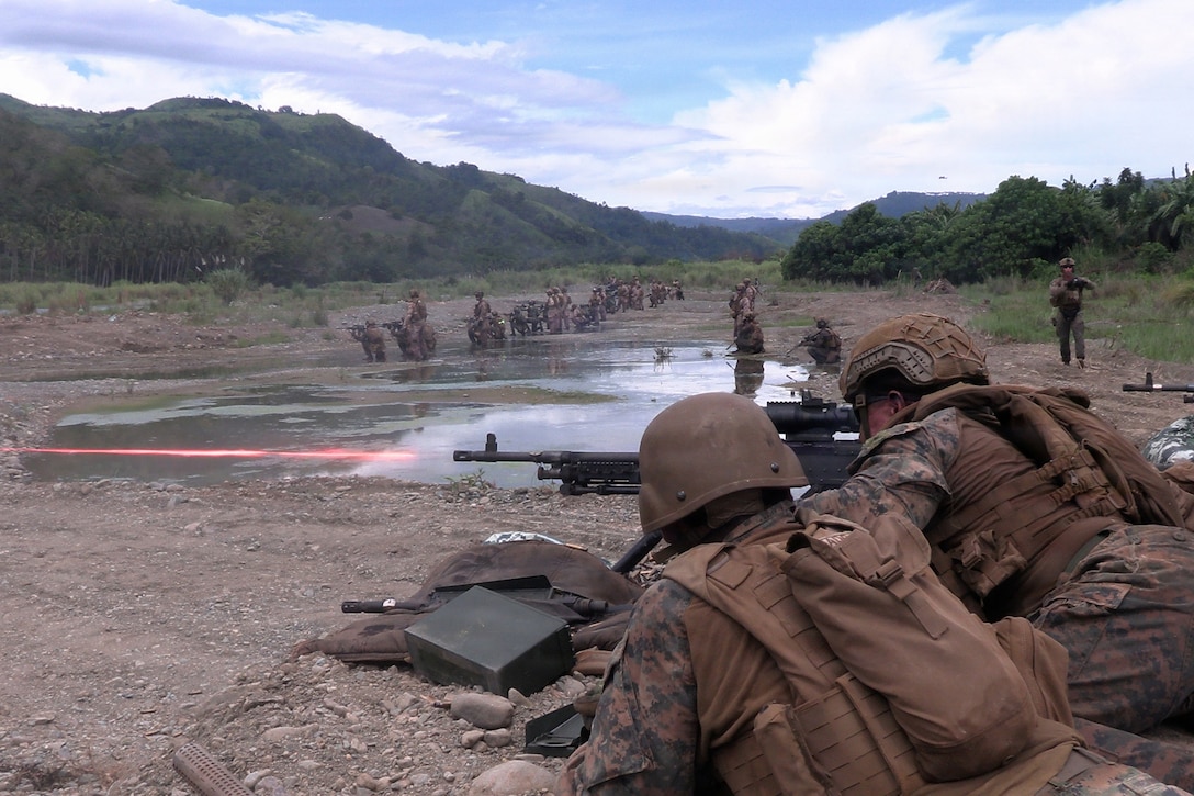 U.S. and Philippine troops fire a weapon in a muddy field, creating a red laser streak as fellow troops train in the distance.