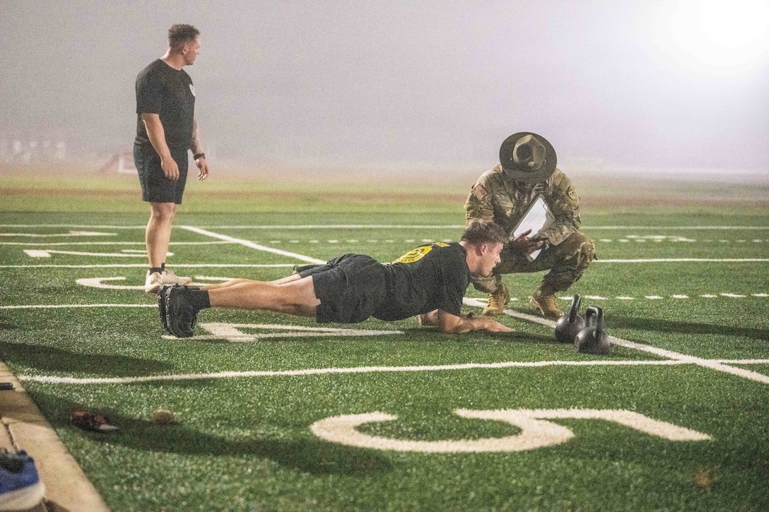 Fog fills the air as a soldier does a plank next to kettle bells on a turf while being evaluated by another soldier as another competitor stands nearby.