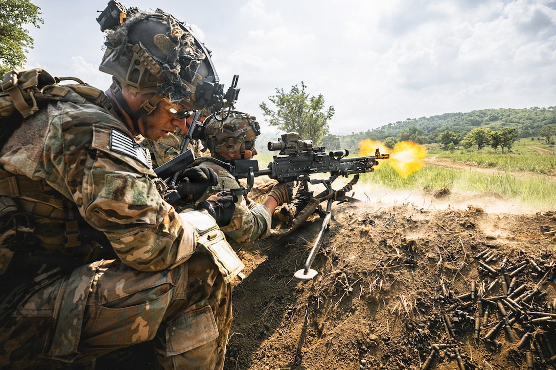 Soldiers in tactical gear fire a weapon from a trench toward a field during the day, creating an orange fireball.