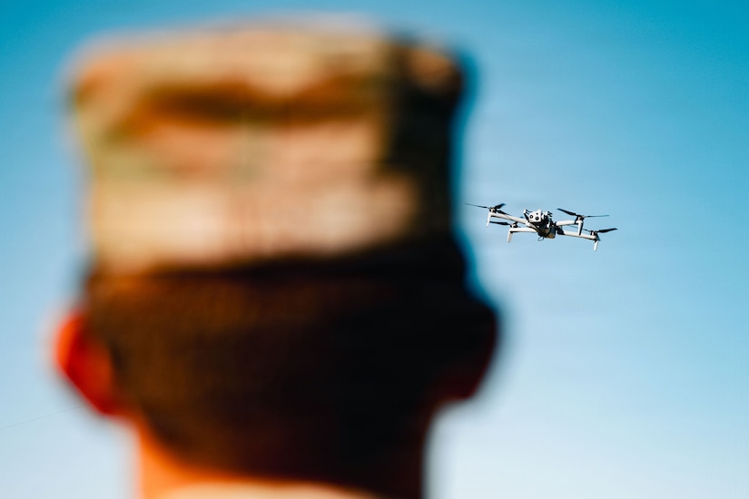 A close-up view of a rear-facing person watching a drone fly through a blue sky overhead.