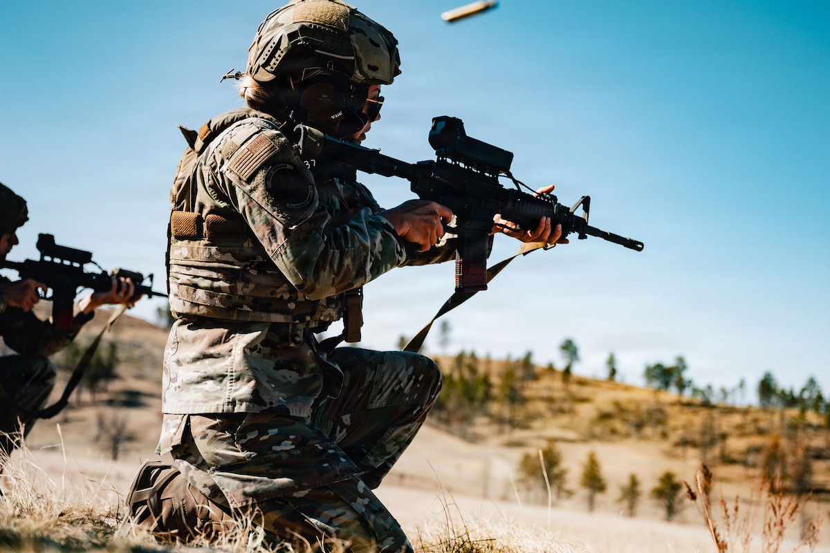 A person wearing a camouflage military uniform and helmet kneels in a grassy terrain and fires a military weapon; an ammunition round disperses out of the weapon.