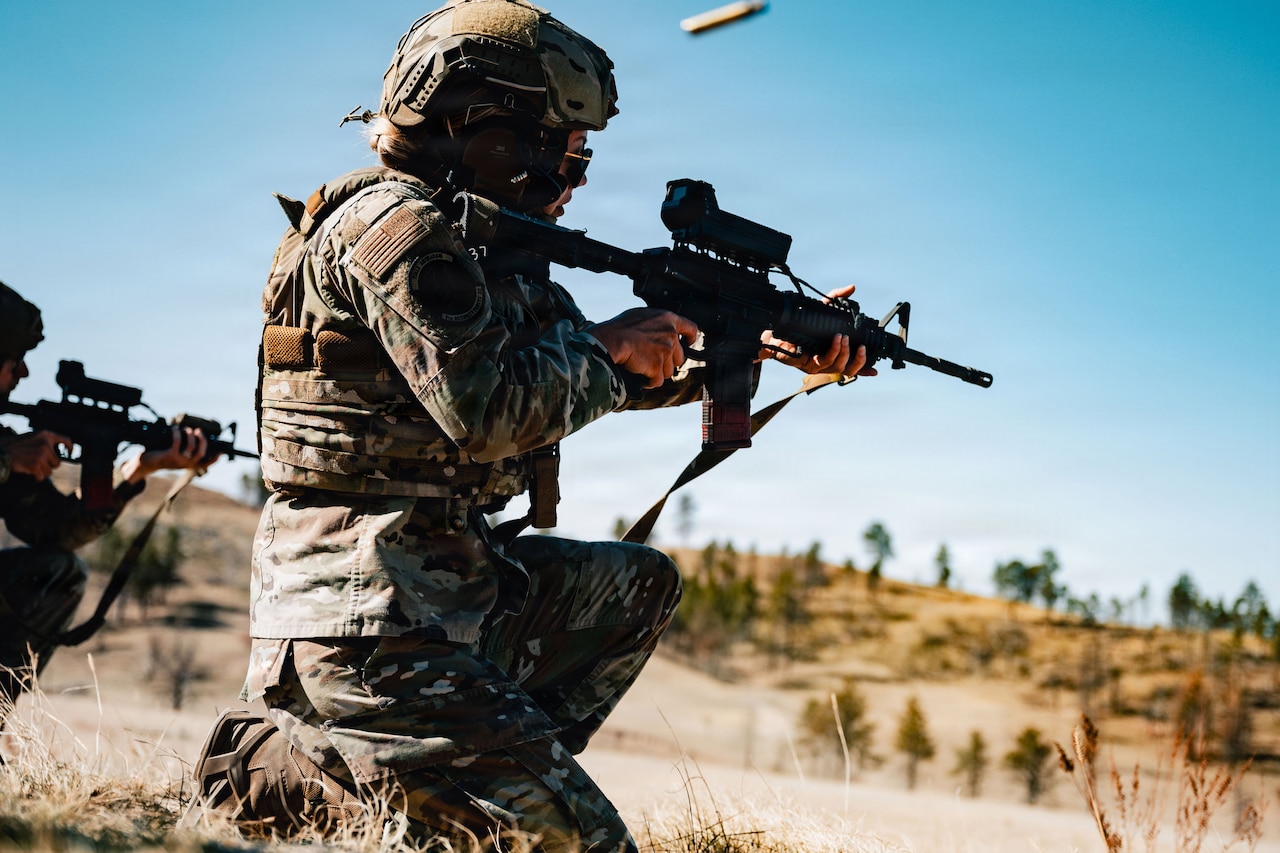 A person wearing a camouflage military uniform and helmet kneels in a grassy terrain and fires a military weapon; an ammunition round disperses out of the weapon.