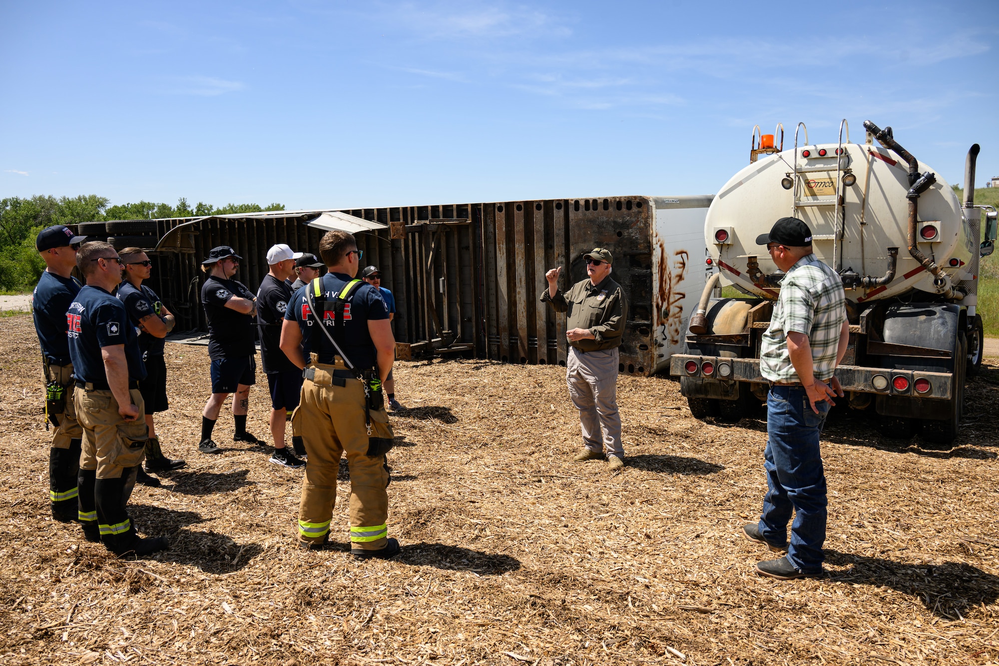 Wayne Francis stands at right center, speaking to firefighters near a large tank truck and an overturned trailer in the background.