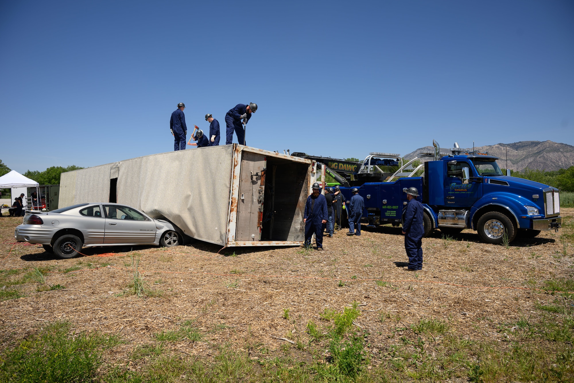 Responders in blue coveralls and hard hats attend to a simulated crash involving an overturned trailer and a car crashed into the trailer, with a tow truck parked in the background.