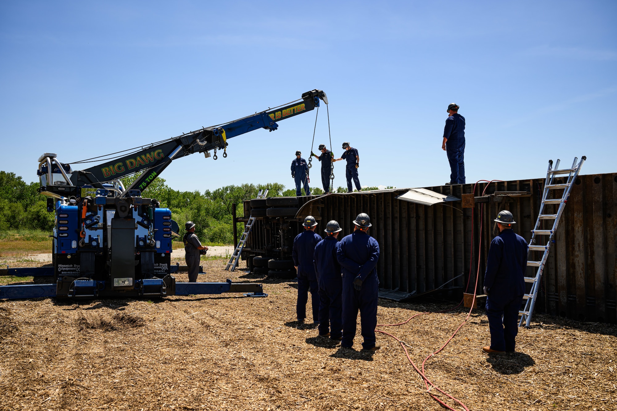 Responders in blue coveralls and hard hats attend to a simulated crash involving an overturned trailer. A crane is parked at left of the trailer with responders scattered through the scene
