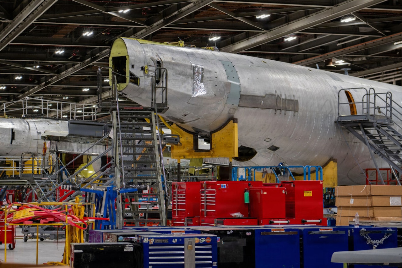 The body of a disassembled aircraft sits in a maintenance hangar surrounded by ladders and large toolboxes. There is a similar aircraft body in the background.