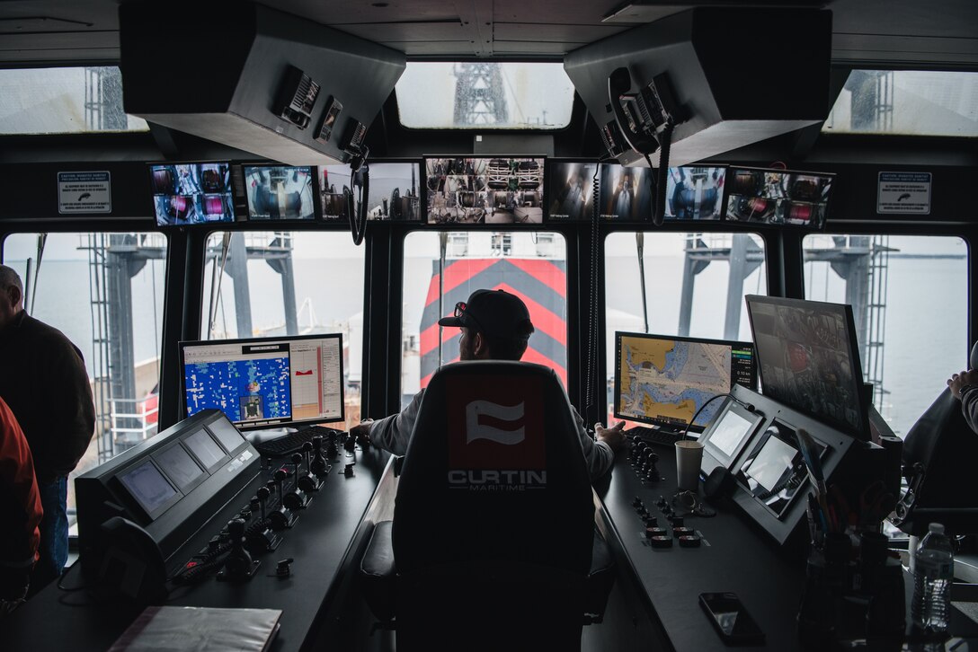 The silhouette of a man wearing a hat and sitting in a control chair as visible against a large wall of windows on a boat. His hands are on control joysticks, operating the machinery and displays that are visible around him.