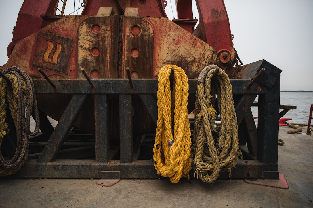 Large yellow ropes are visible bundled along metal hooks in front of a large red dredge bucket on a barge deck. The water and horizon are visible in the background.