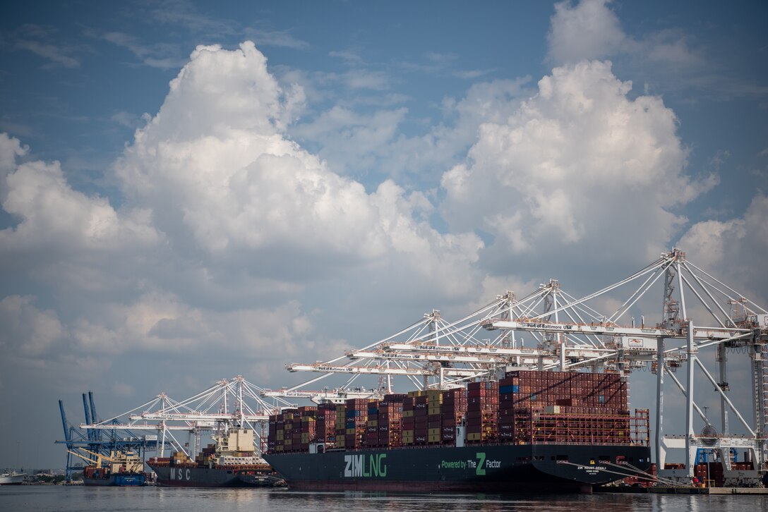 Large white cargo cranes reach out over a long cargo ship and its multicolored containers in a Port.