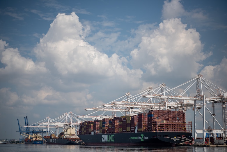 Large white cargo cranes reach out over a long cargo ship and its multicolored containers in a Port.