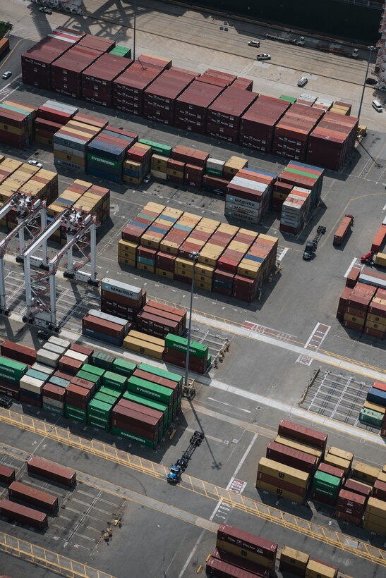 Many multi-colored shipping containers are visible in large stacks as cranes and trucks move them around a large industrial area at the Port of Baltimore.