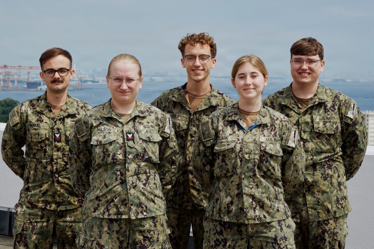 Five people wearing camouflage military uniforms pose for a photo outside with a bay in the background.