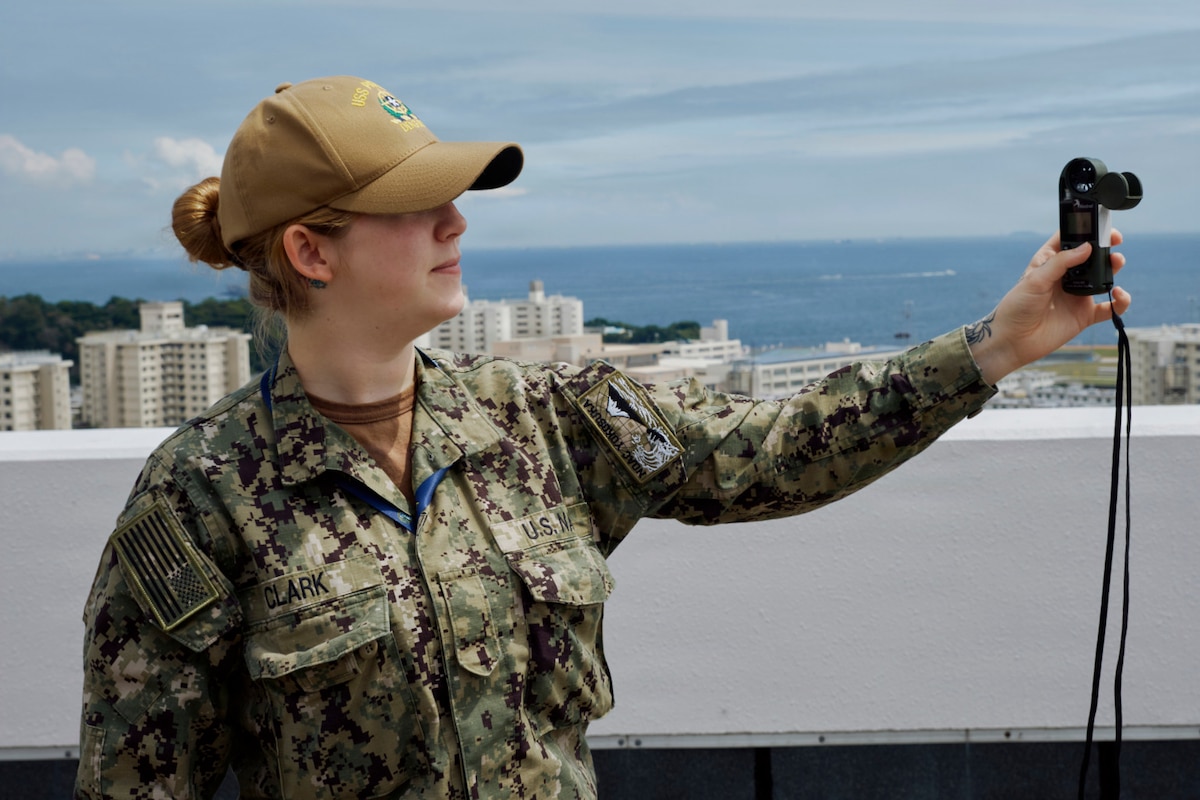 A woman in a military camouflage uniform holds a handheld weather meter in the air outside and looks at the reading.