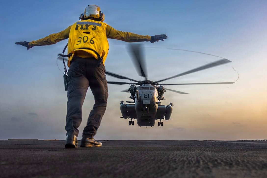 A sailor in a yellow shirt, shown from behind, gestures while standing on a ship's deck to a helicopter hovering just above it.