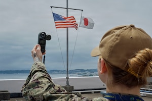 A sailor, shown from behind, holds up a handheld device on the shore as U.S. and Japanese flags wave on a pole nearby.