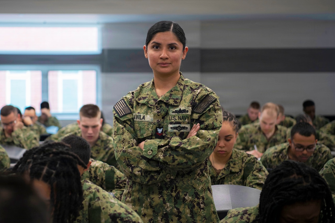 A sailor in a camouflage uniform stands with her arms crossed in a classroom setting, as sailors in similar attire sit at desks around her.