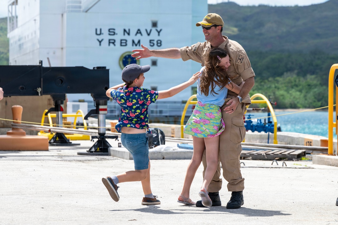 A daughter hugs a sailor as his son runs toward his open arm on a pier with a mountain in the background.