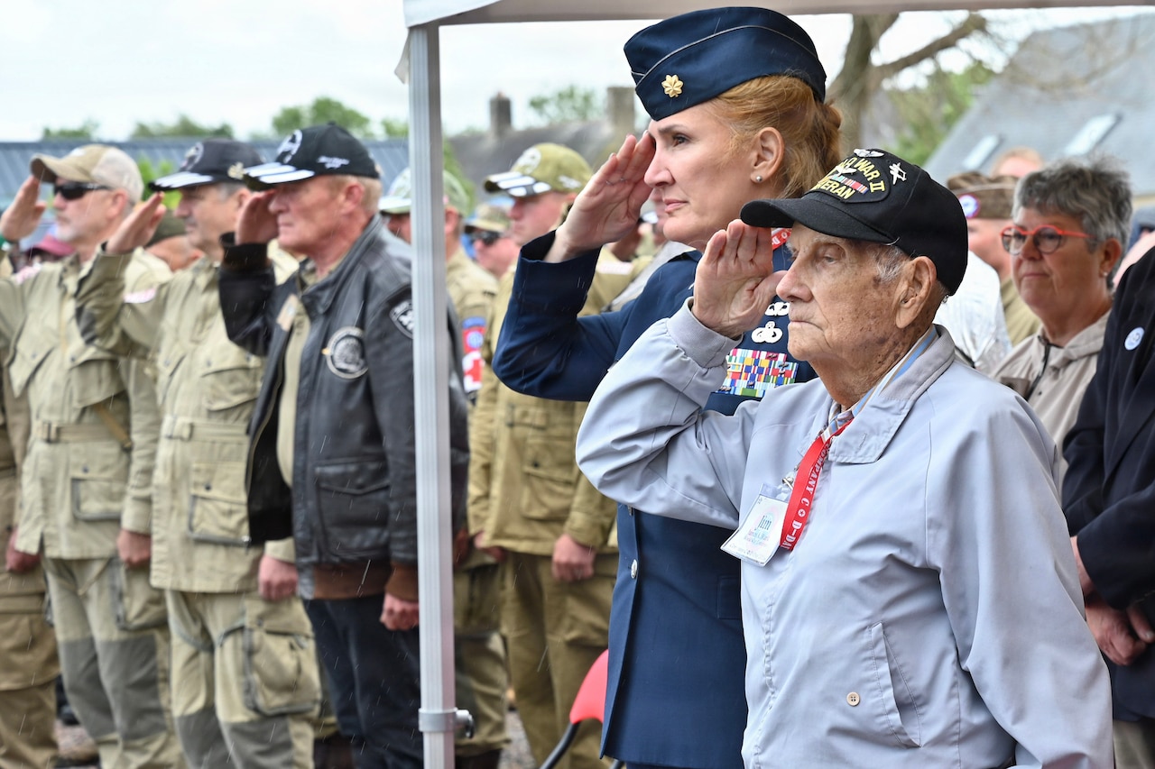 A veteran in a black hat salutes among a standing audience during a ceremony.