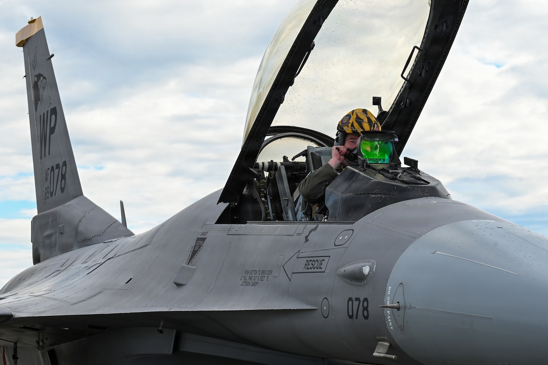 A close-up of an airman gearing up in an open cockpit of a fighter jet against a cloudy, blue sky.