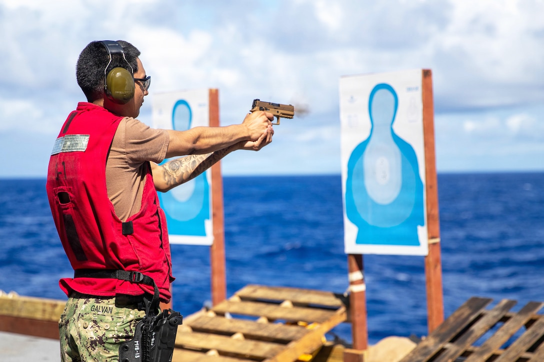 A sailor in a red vest fires a weapon on a ship in blue waters toward a target during the day.