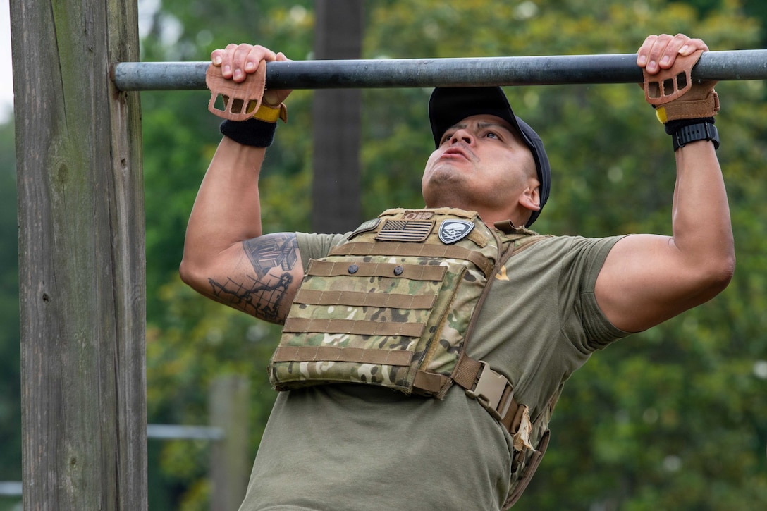 An airman using handgrips and wearing a weighted vest performs a pullup against a wooded background.
