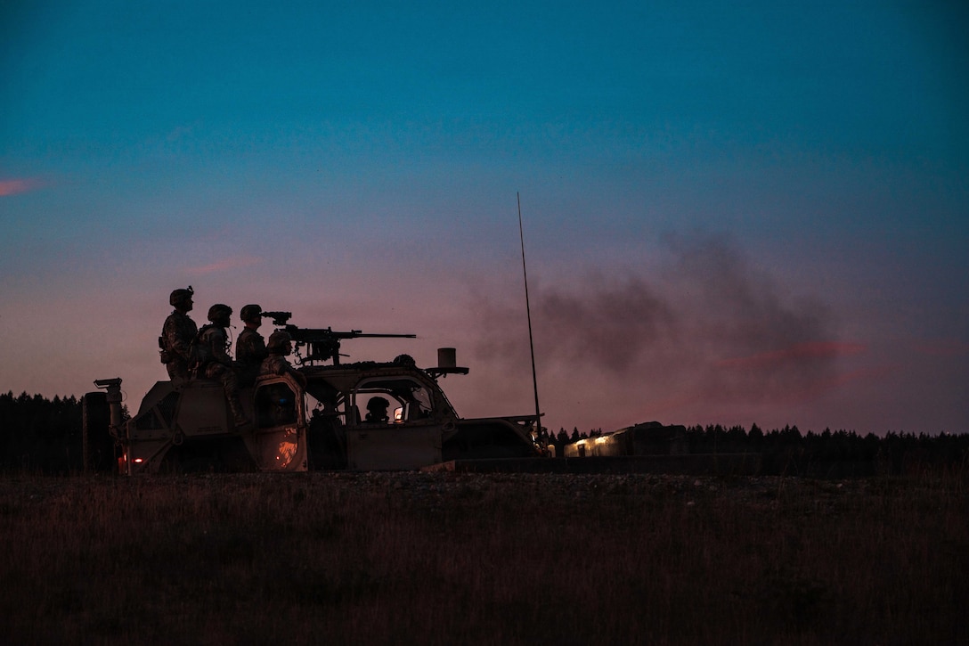 Soldiers fire weapons from a vehicle in dark silhouette in the foreground under pink and blue sky.