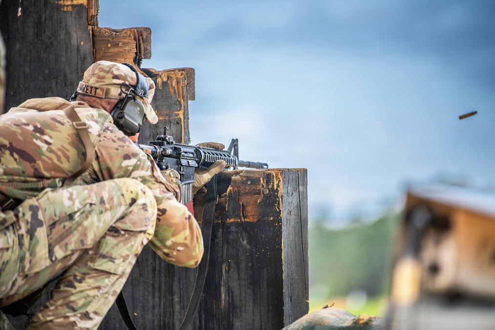A kneeling soldier fires a weapon from a wooden structure. The ammo travels against a blurred blue sky.
