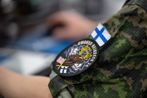 A close-up shot of a Finnish Air Force uniform sleeve, showcasing a distinctive circular patch. The patch prominently displays the text "SMOKED REINDEER 26" curved along its upper and right edges. It features a stylized illustration of a reindeer head with large, elaborate golden antlers, breathing out white smoke or vapor against a dark background. Below the reindeer, two small crossed flags are visible: the United States flag on the left and the Finnish flag on the right. Just above this circular patch, a rectangular patch of the Finnish flag is securely attached to the sleeve. The uniform itself is made of a green, brown, and black camouflage fabric. The background is softly blurred, keeping the focus entirely on the shoulder patches and the uniform's texture.