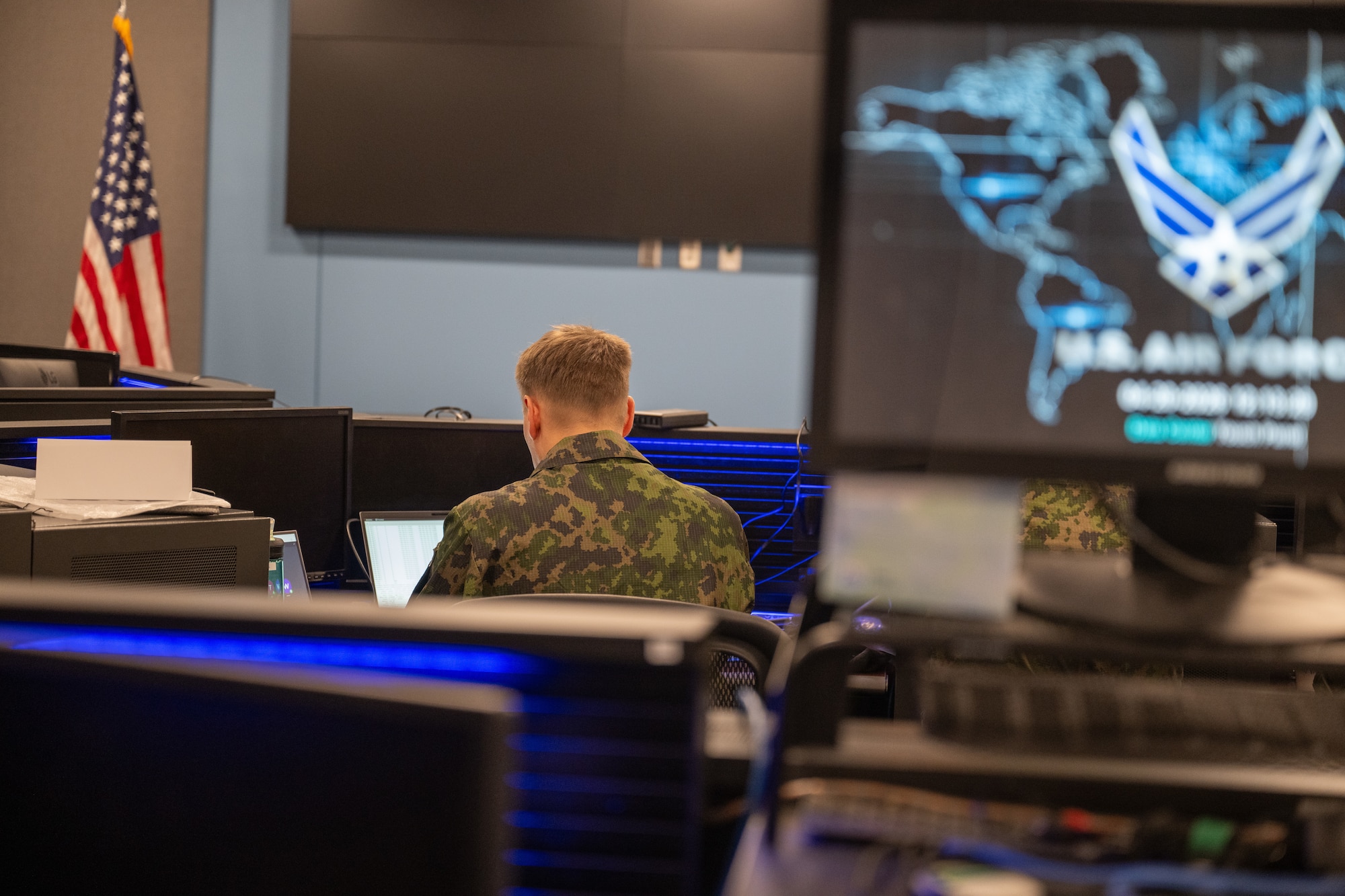 Finnish Air Force member sitting at computer, blurred USAF logo on monitor in foreground with US Flag in background